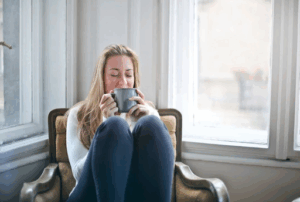 Woman sitting in a chair by the window, drinking a mug with her eyes closed, enjoying overall wellness after her med spa facial treatment in Alameda, CA