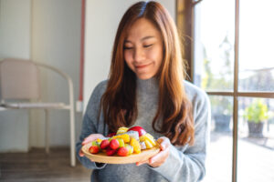 Happy Woman with long brown hair holding a wooden plate of healthy, nutritious fruit standing indoors in her home in Alameda, California near a large window for natural lighting